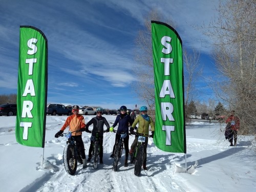 Four cyclists on fat bikes at a snowy start line, flanked by large green "START" banners under a blue sky.