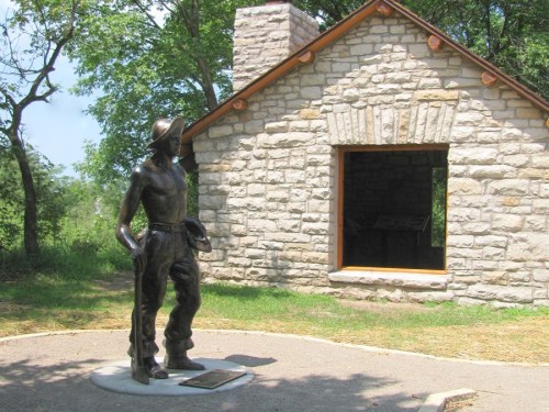 A bronze statue of a man in a hat stands near a stone building surrounded by trees.