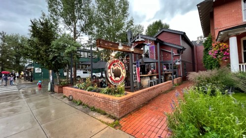 A quaint street scene featuring a rustic building with a sign, surrounded by greenery and a rainy sky.
