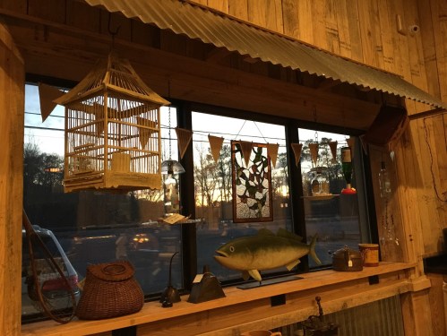 A cozy wooden interior with a large fish decoration, a birdcage, and various hanging ornaments against a sunset backdrop.