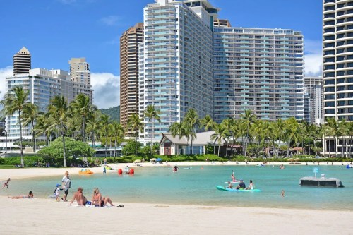 A sunny beach scene with people swimming and kayaking, surrounded by tall buildings and palm trees.
