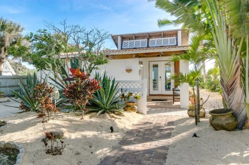 A charming beach house surrounded by tropical plants and sandy pathways under a clear blue sky.