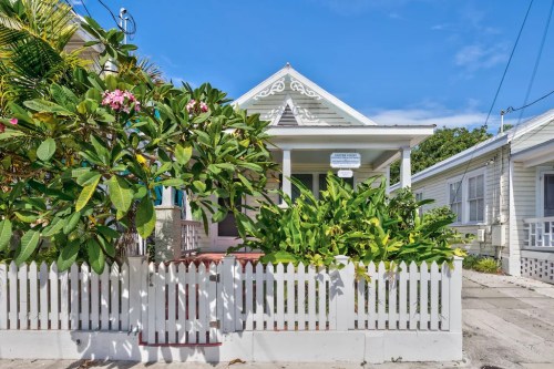 A charming white cottage with a picket fence, surrounded by lush greenery and flowers under a clear blue sky.