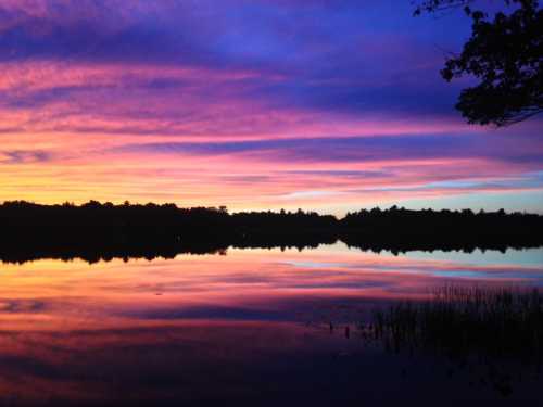 A vibrant sunset reflects on a calm lake, with colorful clouds and silhouettes of trees in the background.