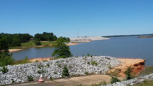 A serene lake view with a rocky shoreline, green trees, and a dam in the distance under a clear blue sky.