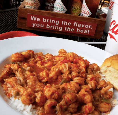 A plate of shrimp and rice in a spicy sauce, with a roll and Tabasco sauce bottles in the background.