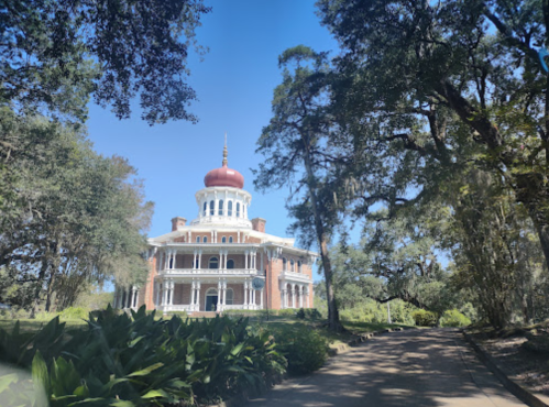 A grand, historic mansion with a red dome, surrounded by lush greenery and tall trees under a clear blue sky.