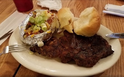 A plate with a grilled steak, a baked potato topped with cheese and green onions, and a soft bread roll.