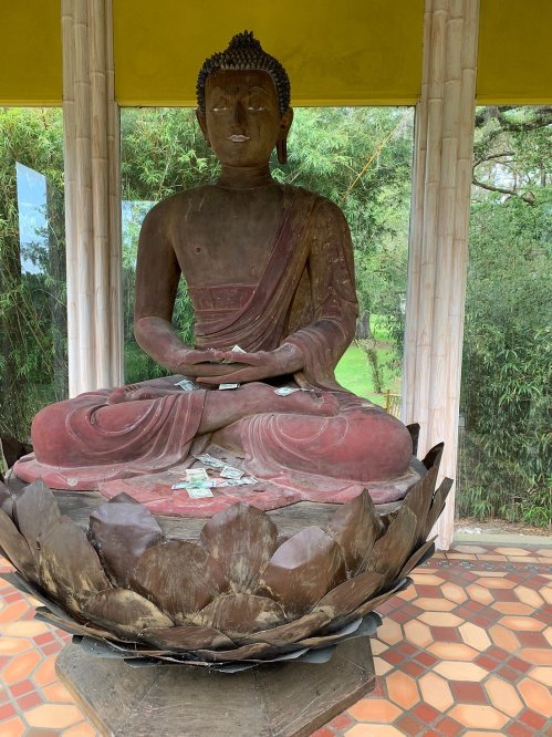 A seated Buddha statue in a lotus position, surrounded by greenery, with offerings placed at its base.