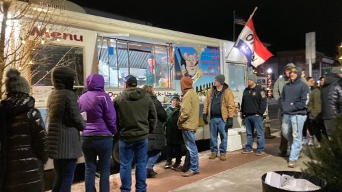 A food truck with a menu sign, surrounded by a line of people waiting outside on a chilly night.