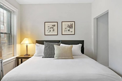 Cozy bedroom with a queen bed, decorative pillows, a lamp, and framed art on the wall. Bright and inviting atmosphere.