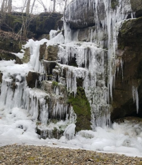 A frozen waterfall with icicles hanging from rocky ledges, surrounded by moss and snow.