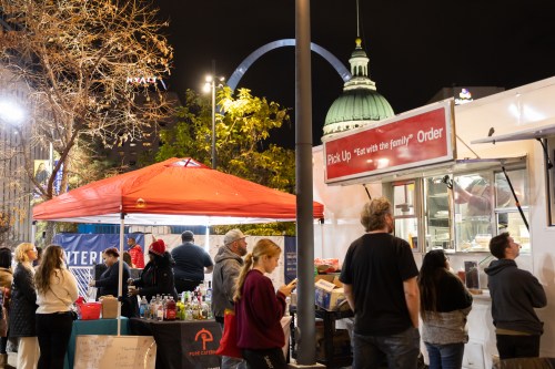 A bustling night market scene with food stalls, people ordering, and a lit-up dome in the background.