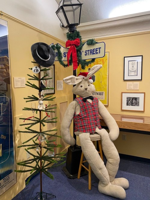 A large stuffed rabbit in a plaid vest sits on a stool next to a decorated Christmas tree and a lamp post.
