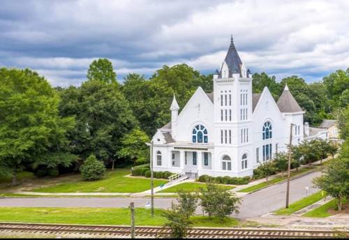 A white church with a tall steeple surrounded by greenery and a railway track nearby under a cloudy sky.