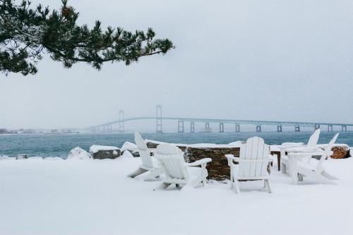 Snow-covered landscape with white Adirondack chairs and a bridge in the background, under a gray winter sky.