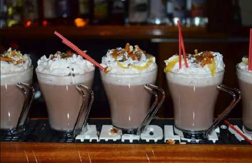 Five frosty drinks topped with whipped cream, chocolate, and nuts, served in clear mugs with red straws.