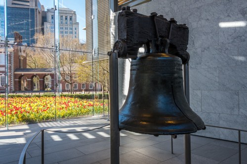 A large bell displayed indoors, with a view of a garden filled with tulips and city buildings in the background.