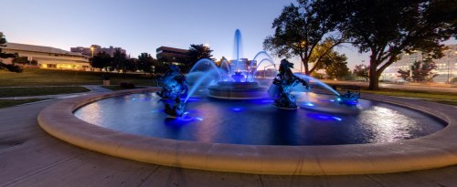 A illuminated fountain with blue lights, featuring sculptures, surrounded by grass and buildings at dusk.