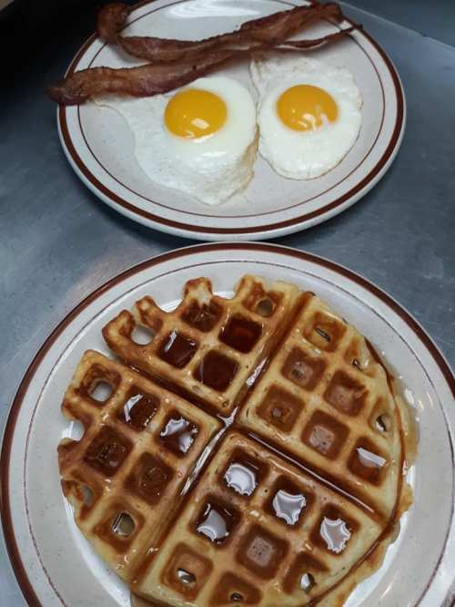 A plate with two sunny-side-up eggs and crispy bacon above a plate of golden waffles drizzled with syrup.