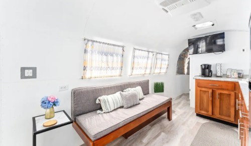 Cozy interior of a modern trailer with a sofa, kitchenette, and large windows, featuring light decor and plants.