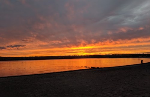 A serene beach at sunset, with vibrant orange and yellow hues reflecting on calm water and a silhouette of a person in the distance.