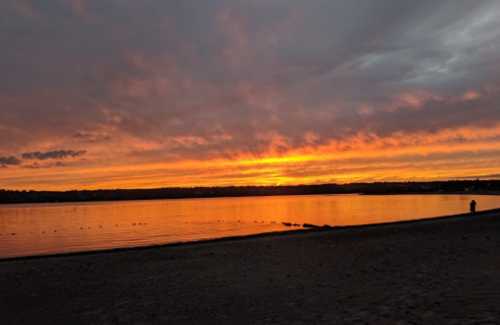 A serene beach at sunset, with vibrant orange and yellow hues reflecting on calm water and a silhouette of a person in the distance.