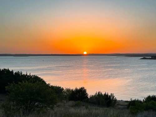 A serene sunset over a calm lake, with vibrant orange and yellow hues reflecting on the water. Silhouetted trees in the foreground.