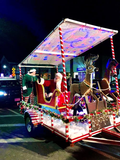 A festive float featuring Santa, an elf, and reindeer decorations, illuminated with colorful lights at night.