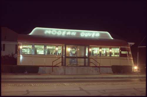 A vintage diner at night, illuminated with neon lights, featuring a curved roof and large front windows.