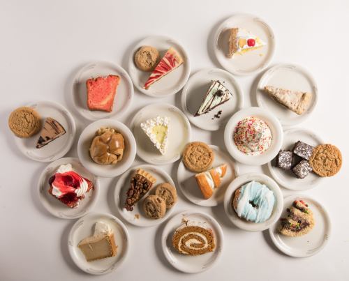 A variety of desserts on small plates, including cakes, cookies, and pastries, arranged on a white surface.