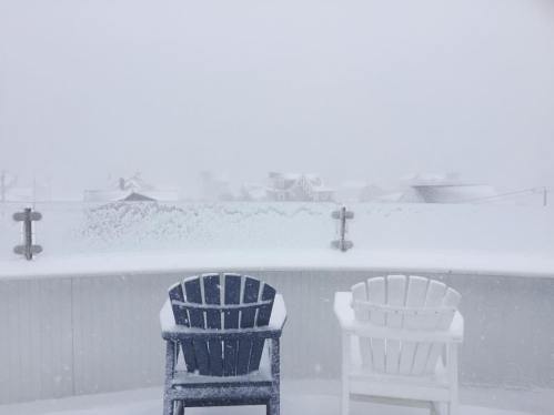 Two empty chairs on a snowy balcony, with heavy snowfall obscuring distant houses and a gray sky.
