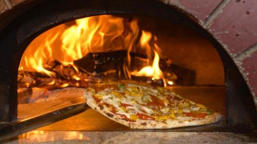 A freshly baked pizza being pulled from a wood-fired oven with flames in the background.