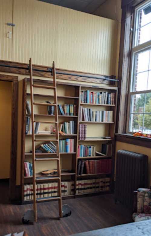 A wooden ladder beside a tall bookshelf filled with books, in a cozy room with large windows and wooden accents.