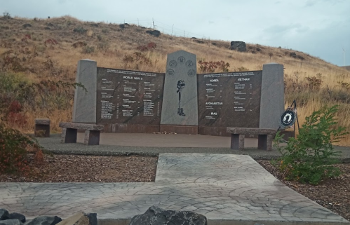 A memorial with stone panels listing wars and names, set against a dry, grassy hillside under a cloudy sky.