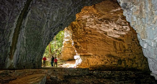 Three people stand inside a large cave, illuminated by natural light, surrounded by rocky walls and lush greenery outside.
