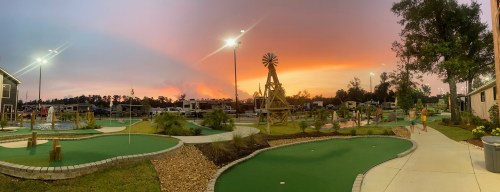 A scenic mini-golf course at sunset, featuring a windmill and vibrant sky with orange and purple hues.