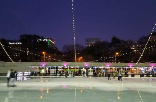 A skating rink at dusk, adorned with lights, with people skating and a building in the background.