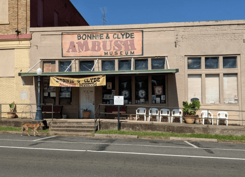 Exterior of the Bonnie & Clyde Ambush Museum, featuring a sign and empty chairs outside, with a dog walking by.