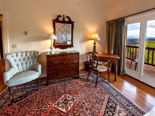Cozy room featuring a blue armchair, wooden dresser, desk, and large windows with scenic views.