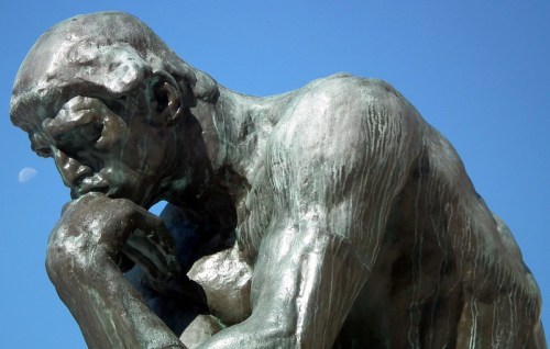 A close-up of a bronze sculpture of a muscular figure deep in thought against a clear blue sky.