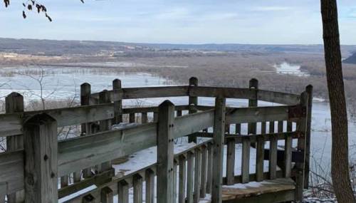 A wooden observation deck overlooks a river valley with trees and hills in the background under a clear sky.