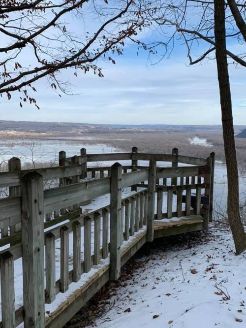 A wooden overlook with a railing, surrounded by snow, offers a view of a river and distant hills under a cloudy sky.