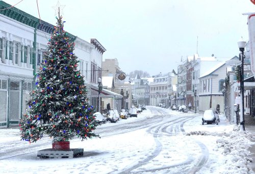A snowy street scene featuring a decorated Christmas tree and snow-covered buildings.