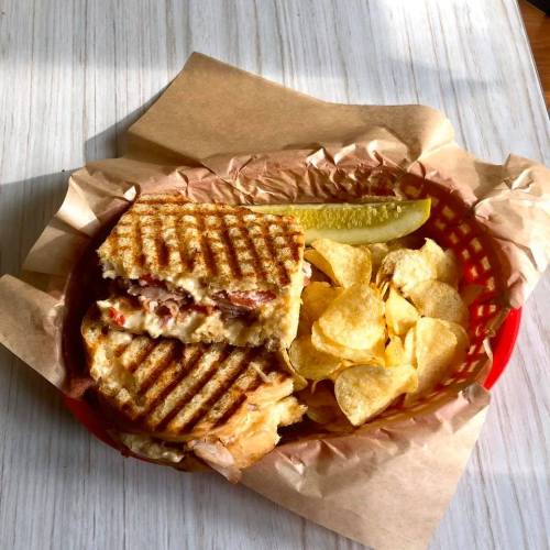 A grilled sandwich with layers of filling, served with potato chips and a pickle slice in a red basket.