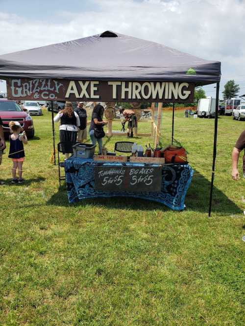 A tent for Grizzly Axe Throwing with a sign, axes for sale, and people participating in the activity on a grassy field.