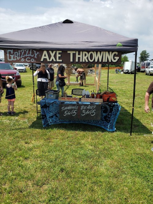 A tent for Grizzly Axe Throwing with a sign, axes for sale, and people participating in the activity on a grassy field.