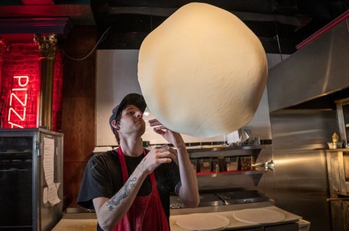 A chef in a black shirt and red apron tosses a large ball of pizza dough in a pizzeria kitchen.