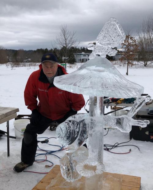 A man in a red jacket poses beside a detailed ice sculpture in a snowy landscape.