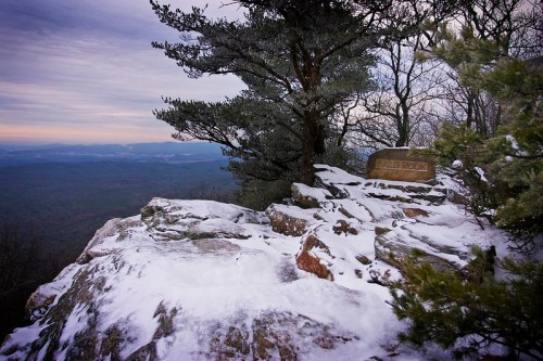 A snowy rocky outcrop with a tree, overlooking a vast mountainous landscape under a cloudy sky.